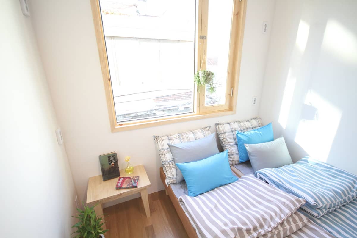 A small, bright room with a bed, a bedside table, a window, and a potted plant.
