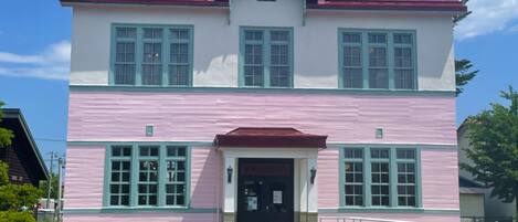 A two-story building with a red roof and pink walls, featuring a central entrance with steps and a small porch.