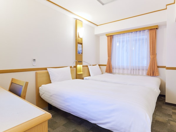 A hotel room with two beds, a desk, a bedside table, a lamp, a window with curtains, and a wall-mounted phone.