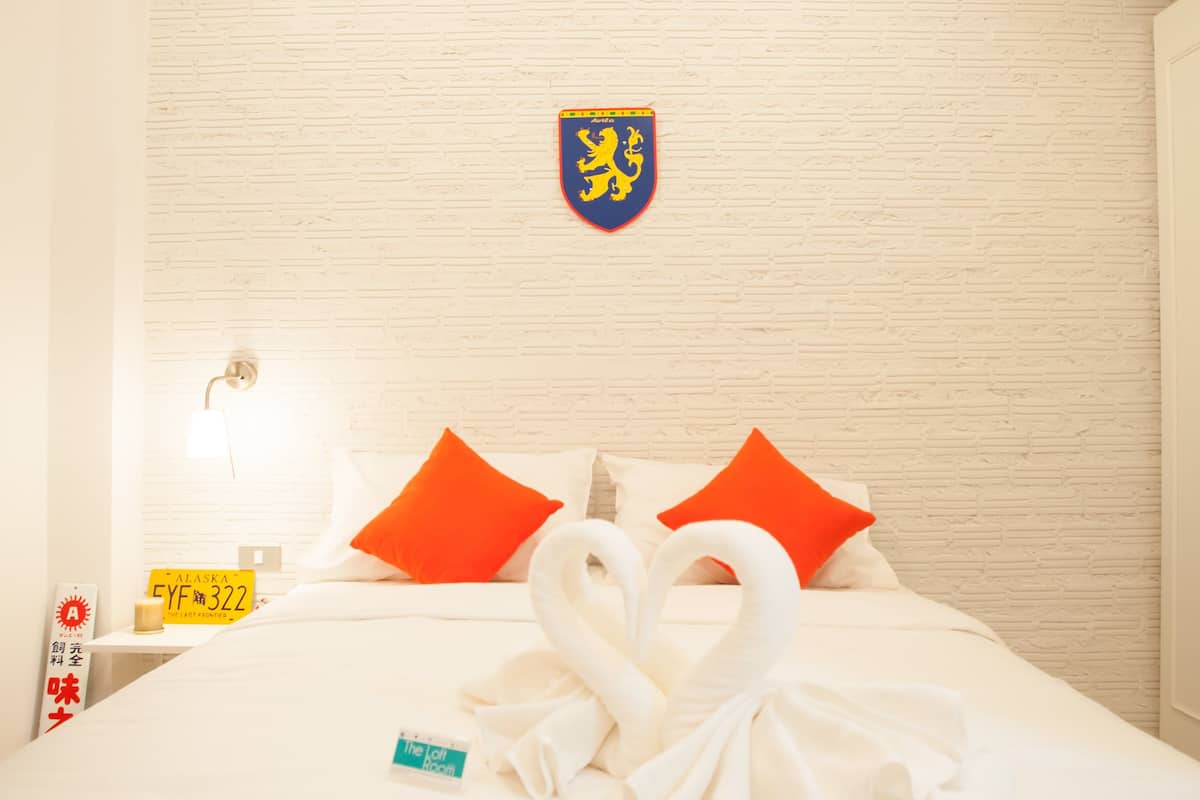 A hotel room with a white brick wall, a bed with white bedding and orange pillows, a towel swan decoration, a coat of arms on the wall, and a nightstand with a lamp and a sign.