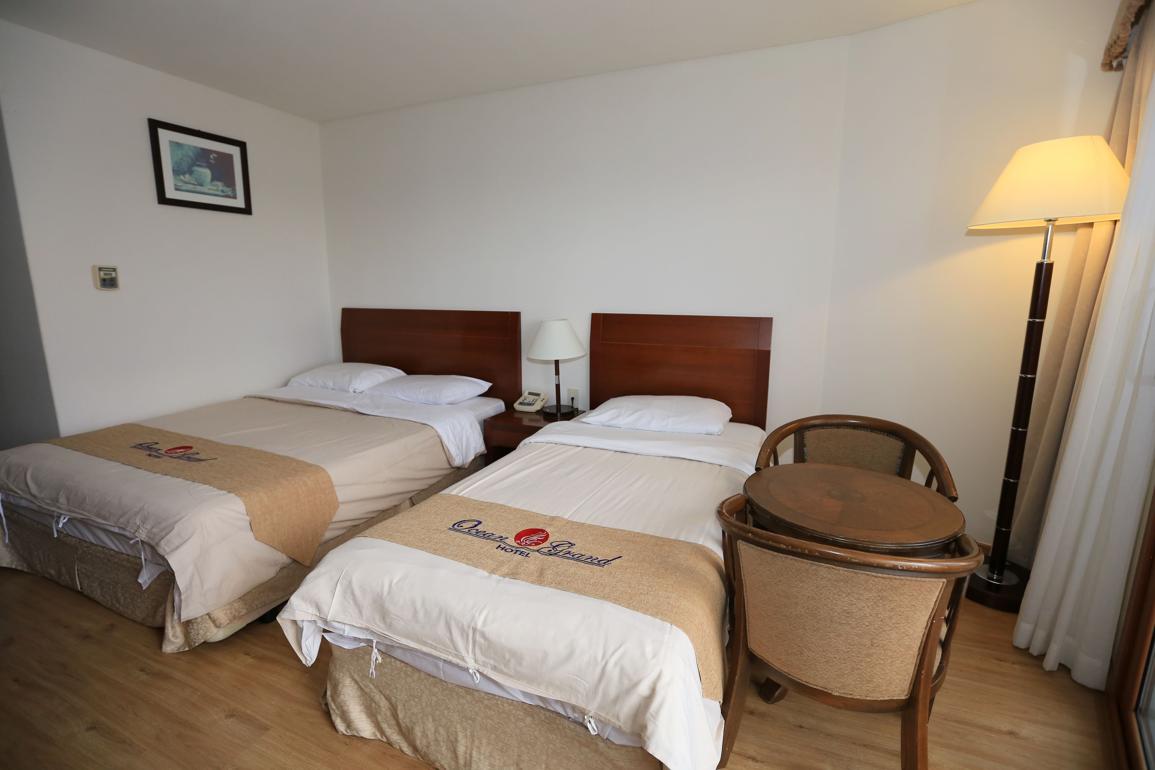 Una habitación de hotel con dos camas, una silla de madera, una mesita de noche con lámpara y un cuadro en la pared.