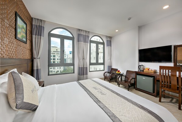 A hotel room with a bed, two windows, a desk with a television, and a small refrigerator.