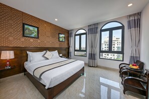 A hotel room with a large bed, wooden headboard, two framed pictures, a nightstand with a lamp, a dining area with a fruit bowl, and large windows with curtains.