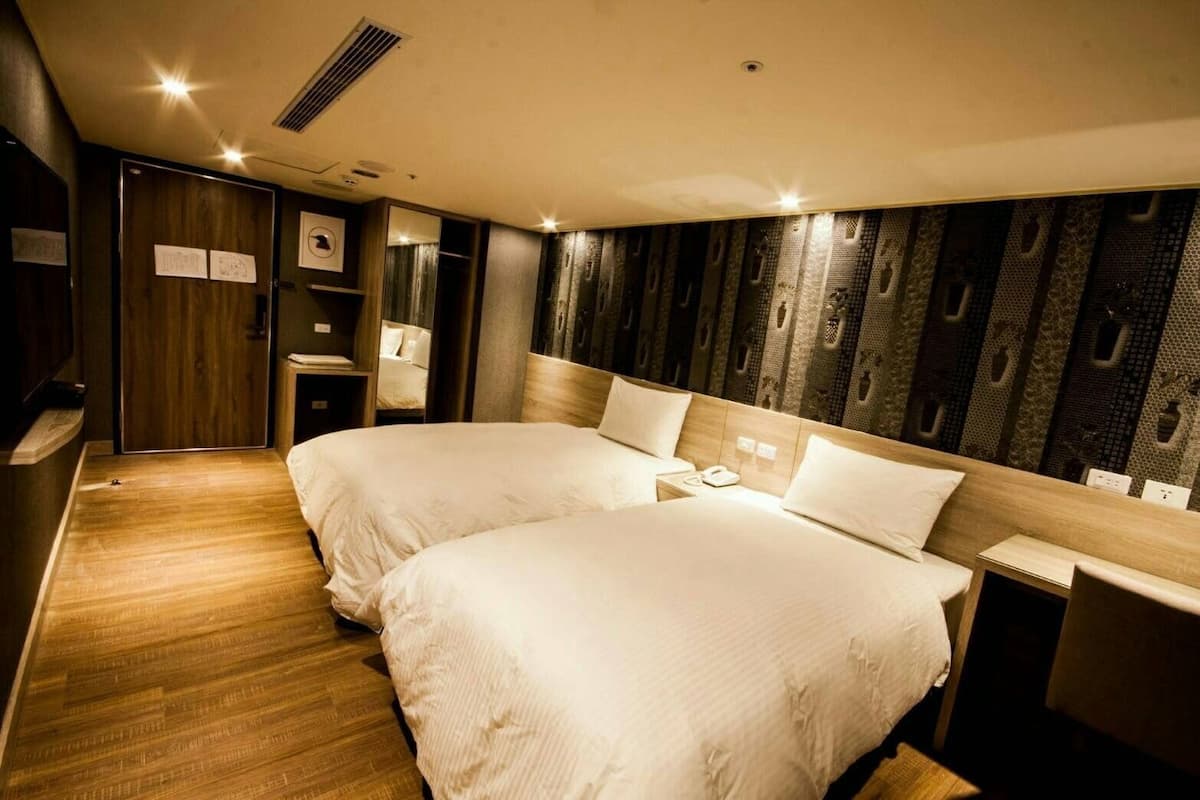 A hotel room with two beds, a wooden floor, a headboard with a patterned design, and a small bedside table with a lamp.