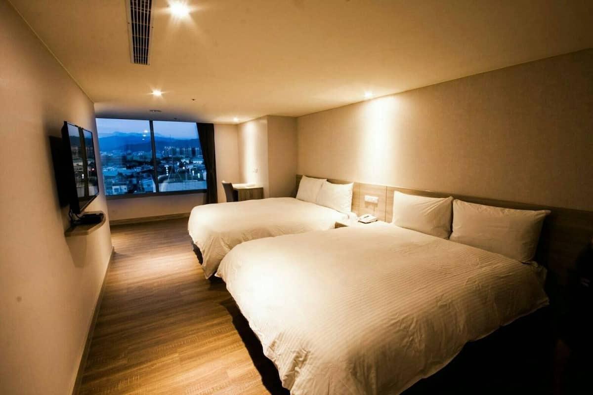 A hotel room with two beds, a TV, a desk, and a large window with a city view.