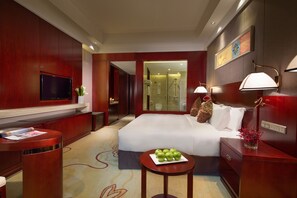 Una habitación de hotel con una cama grande, un televisor, un escritorio con lámpara y una mesita con manzanas.