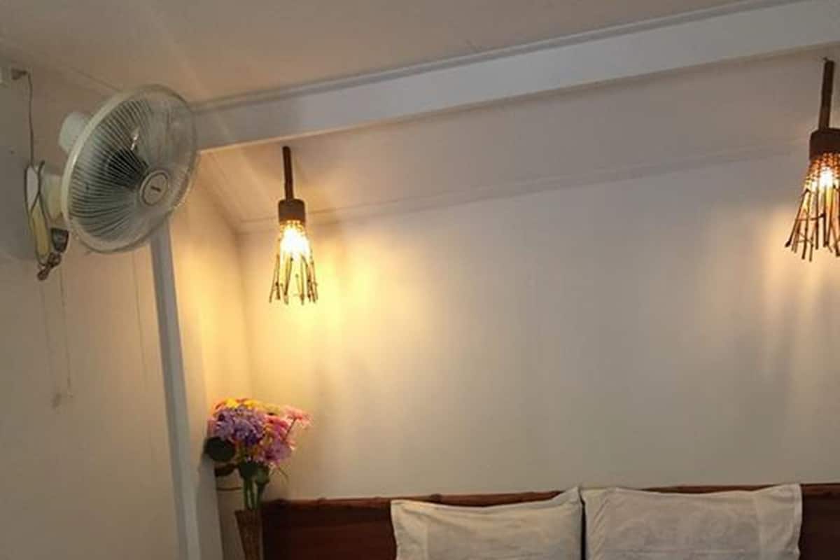 Ein Einzelbett mit weißer Matratze und zwei Kissen, ein Nachttisch mit Blumenvase, ein Wandventilator und zwei Pendelleuchten.
