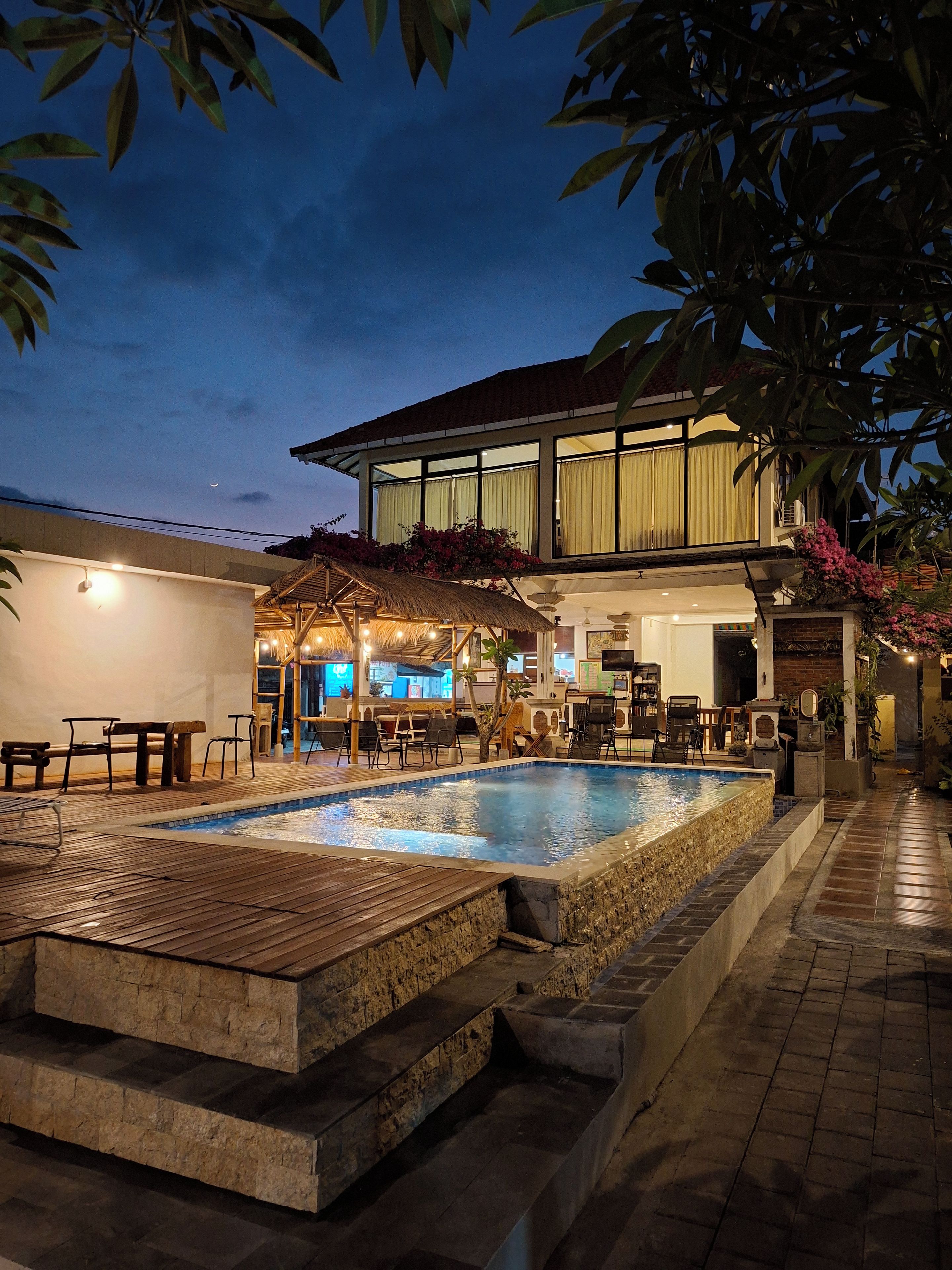 A swimming pool with a wooden deck and stone steps, surrounded by outdoor seating and a building with large windows.