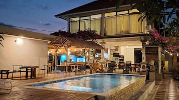 Una piscina con cubierta de madera y escalones de piedra, rodeada de mobiliario exterior y un edificio con amplias ventanas.