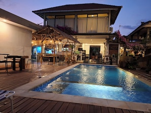 A swimming pool with a wooden deck, a covered seating area, and a building with a tiled roof.