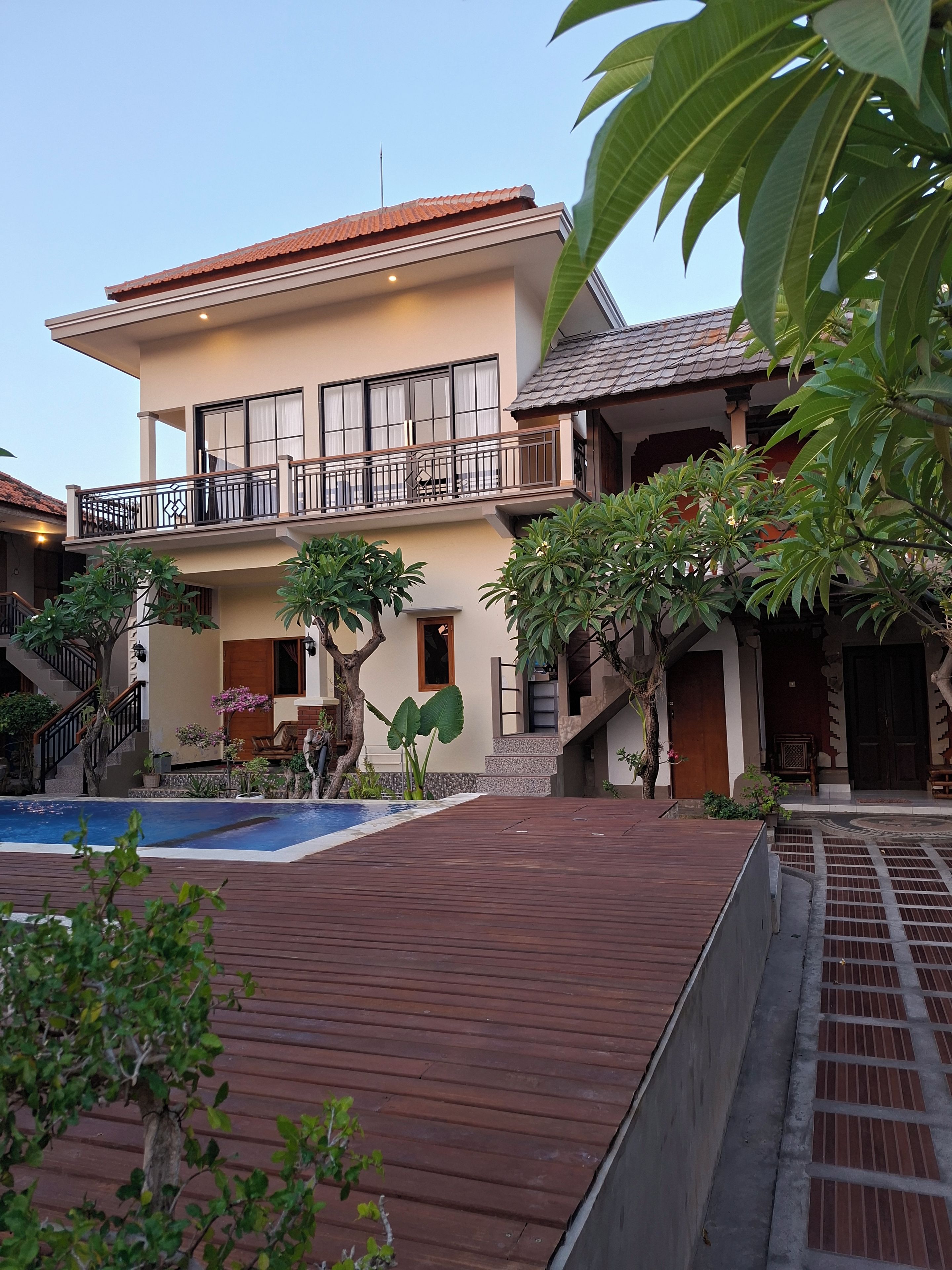 Un edificio de dos pisos con piscina, muelle de madera y plantas tropicales.