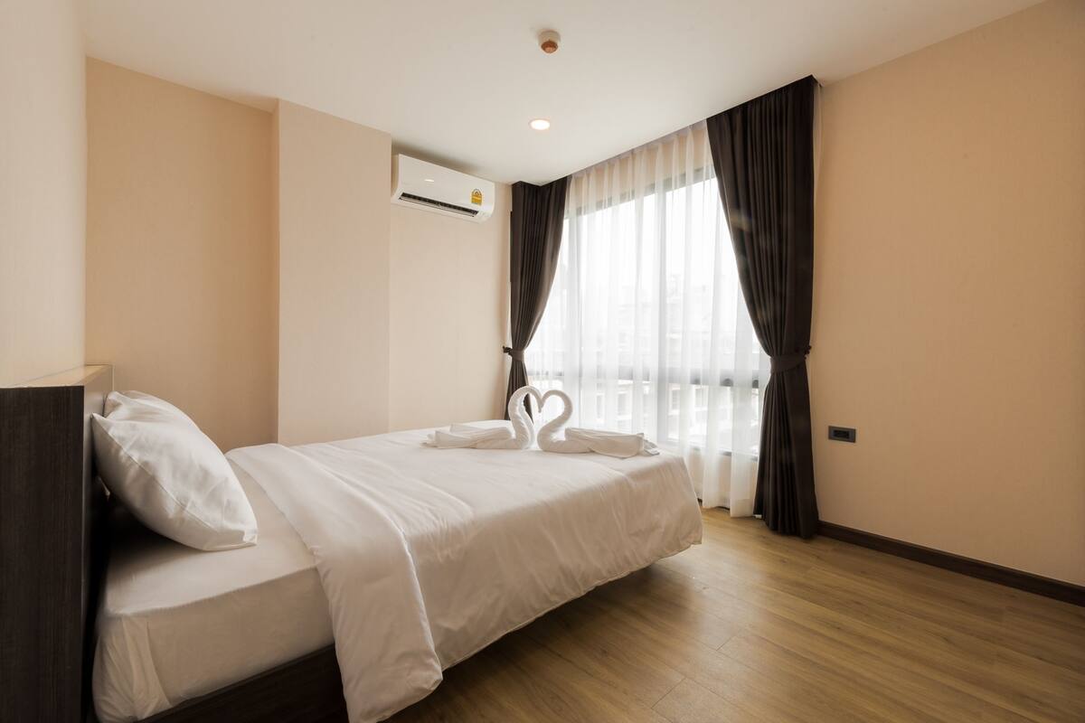 Une chambre d’hôtel avec un lit, un oreiller, une serviette blanche, une décoration en forme de cœur faite avec des serviettes, une fenêtre avec des rideaux et un appareil de climatisation.