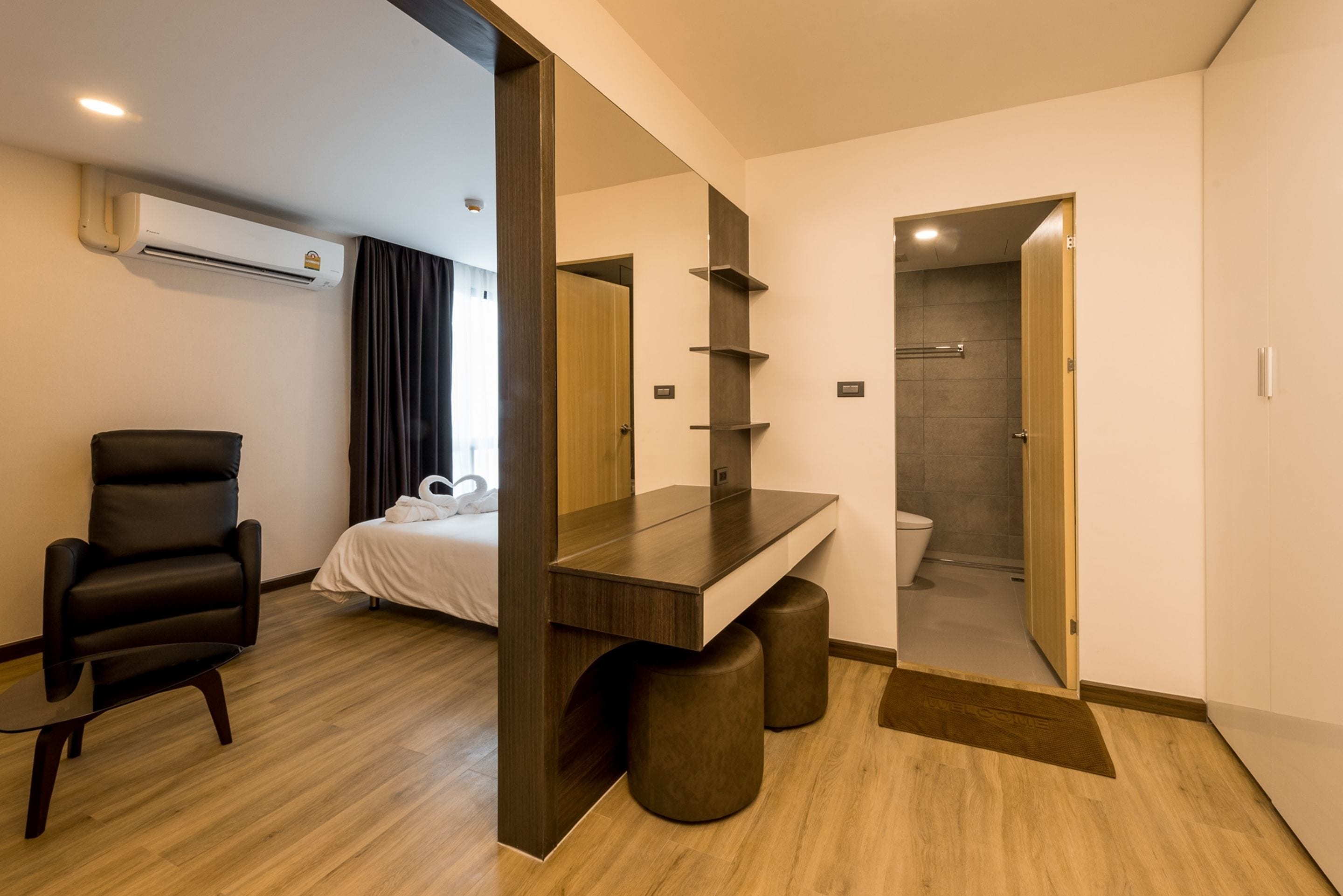 Una habitación de hotel moderna con suelo de madera, un sillón de cuero negro, una cama con ropa blanca, un escritorio de madera con espejo y una zona de ducha visible a través de una puerta abierta.