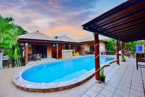 A swimming pool with a tiled deck, a covered seating area, and a building with a tiled roof. - Panisara Pool Villa (Hua Hin)
