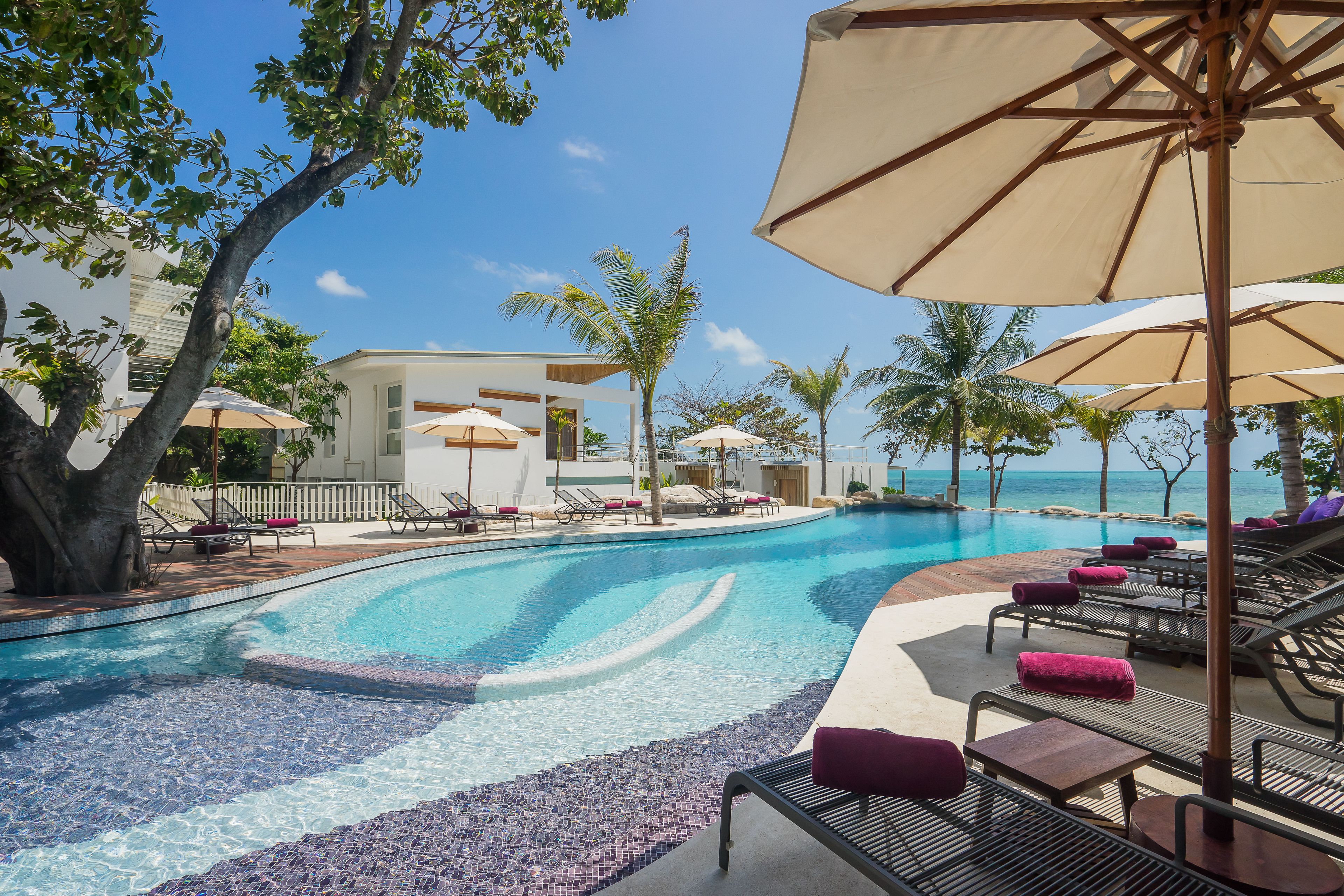 A pool area with lounge chairs and umbrellas, surrounded by palm trees and a clear view of the ocean.