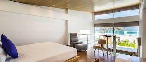A modern hotel room with a large bed, a seating area with a table, and a balcony with a view of the sea.