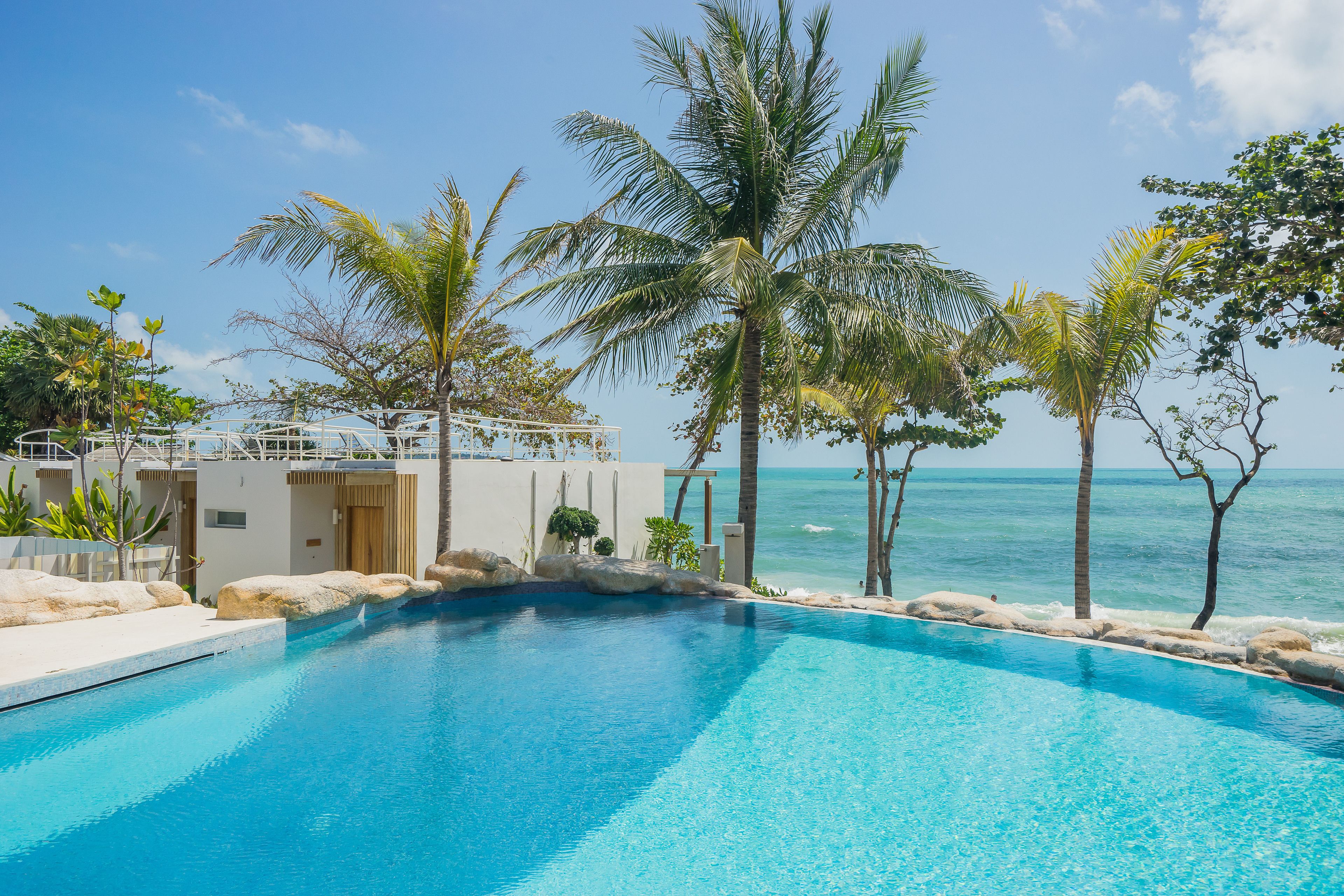A clear blue pool with a view of the ocean, surrounded by palm trees and a white building with a balcony.