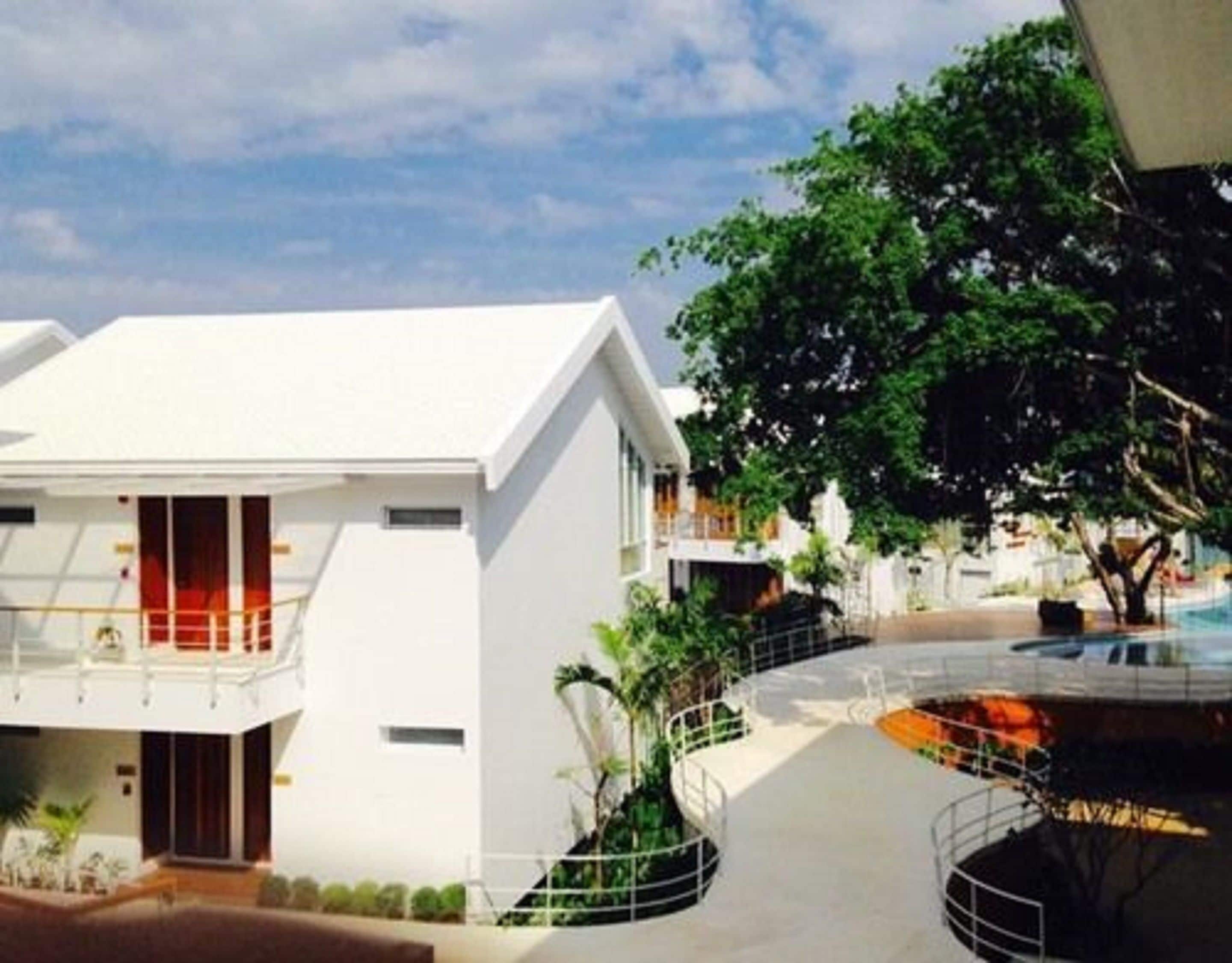 Un edificio moderno con techo plano, ventanas grandes y un balcón. Cerca hay un árbol y una pequeña fuente de agua.