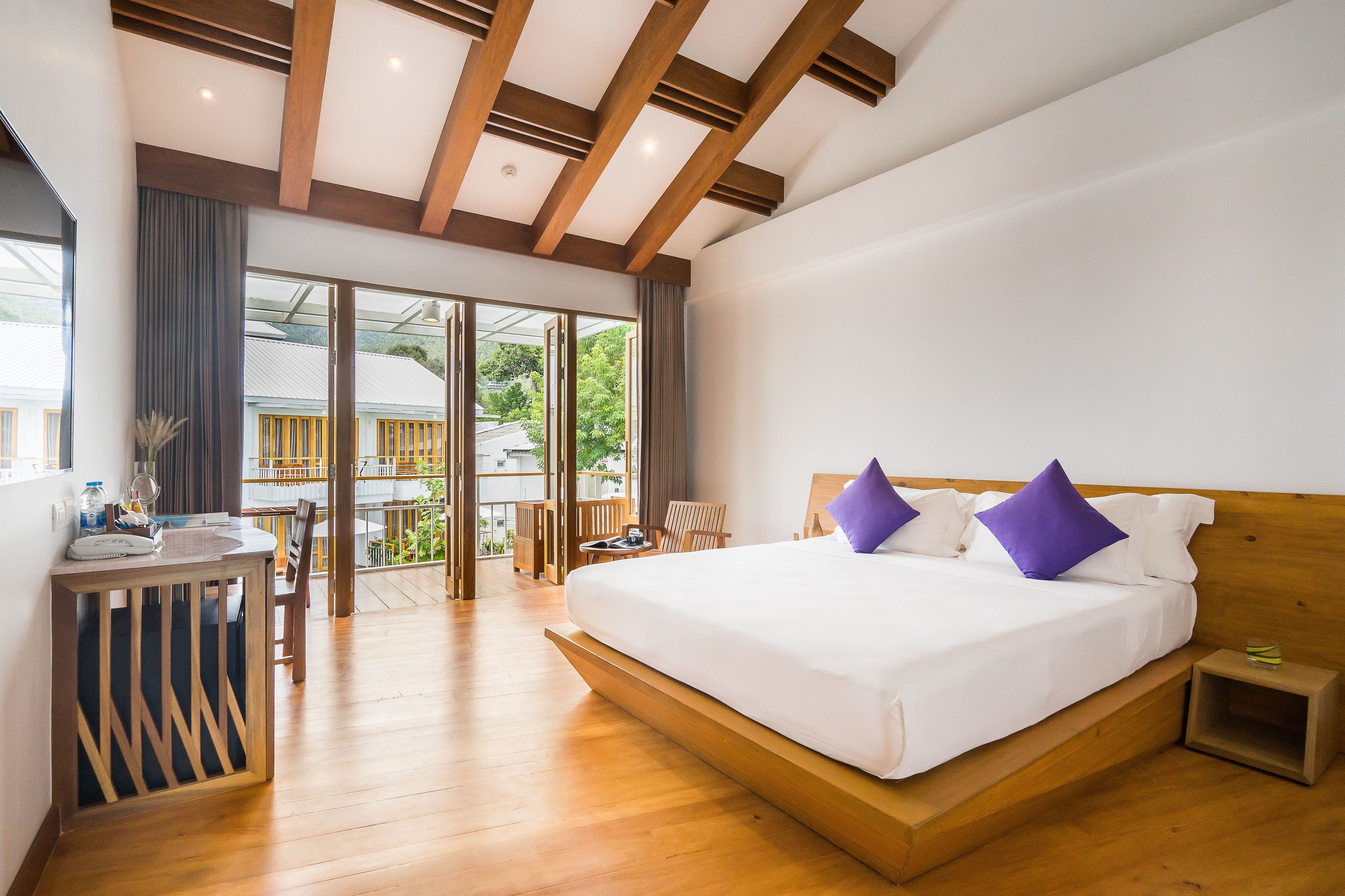 A modern hotel room with a large bed, wooden ceiling beams, a balcony with a view, and a small table with a chair.