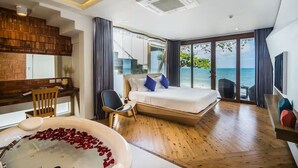 A modern hotel room with a wooden floor, a large bed with blue pillows, a desk with a chair, a bathtub with rose petals, and a view of the sea through large windows. - White Sand Samui Resort (Koh Samui)