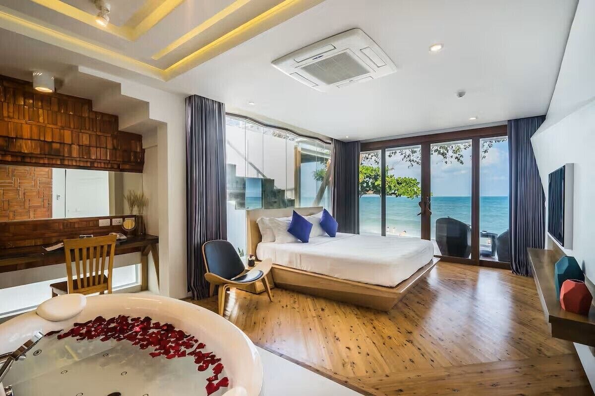 Una habitación de hotel moderna con suelo de madera, una cama grande con almohadas azules, un escritorio con una silla, una bañera con pétalos de rosa y vistas al mar a través de amplios ventanales.