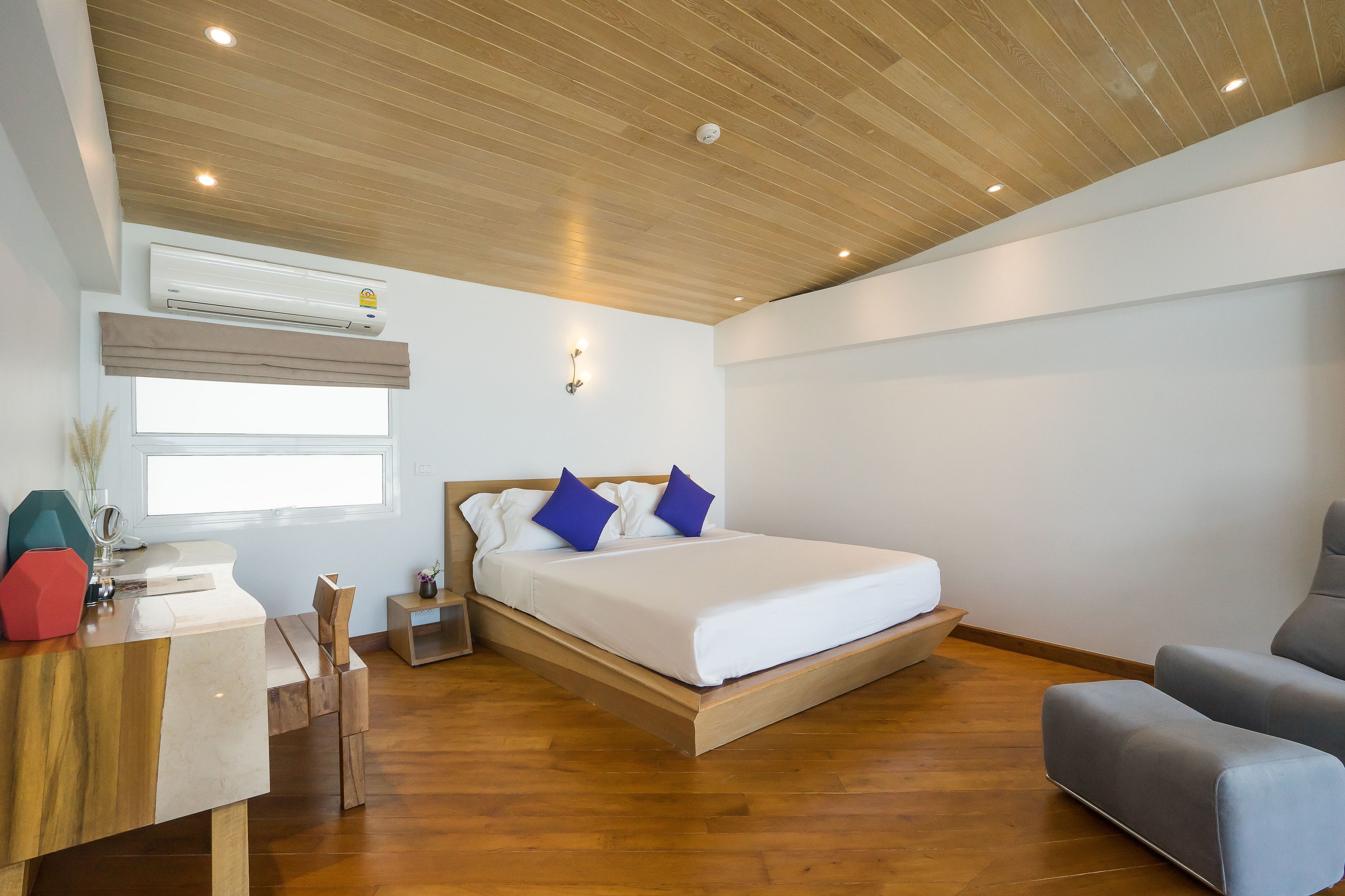 Un dormitorio moderno con techo de madera, una cama con ropa de cama blanca y almohadas azules, un escritorio con un florero y un sillón gris.