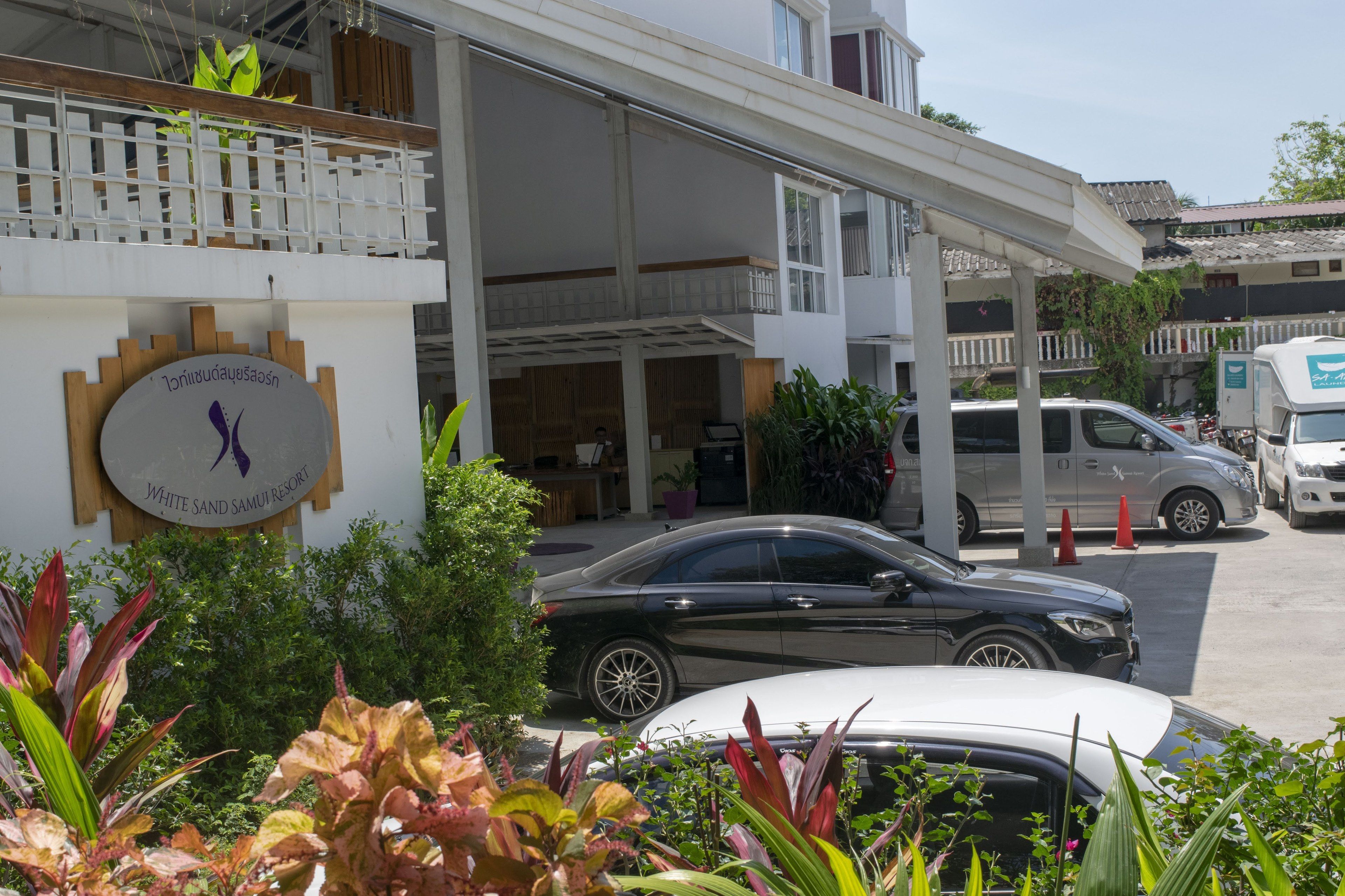 A hotel entrance with a sign for White Sand Samui Resort, a covered parking area, and a white car parked in front.