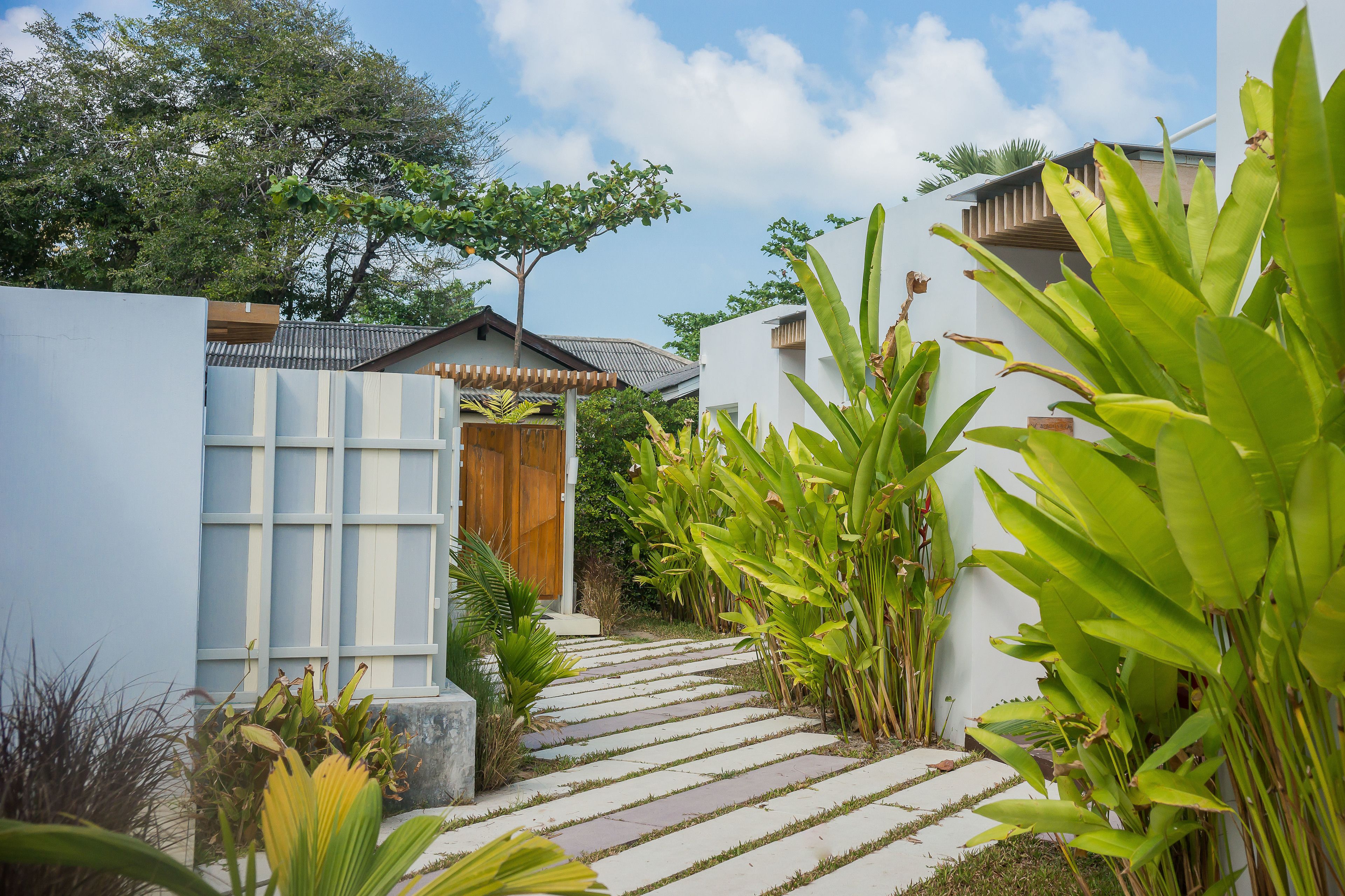 A pathway leading to a house with a wooden door, surrounded by lush greenery and a clear sky.