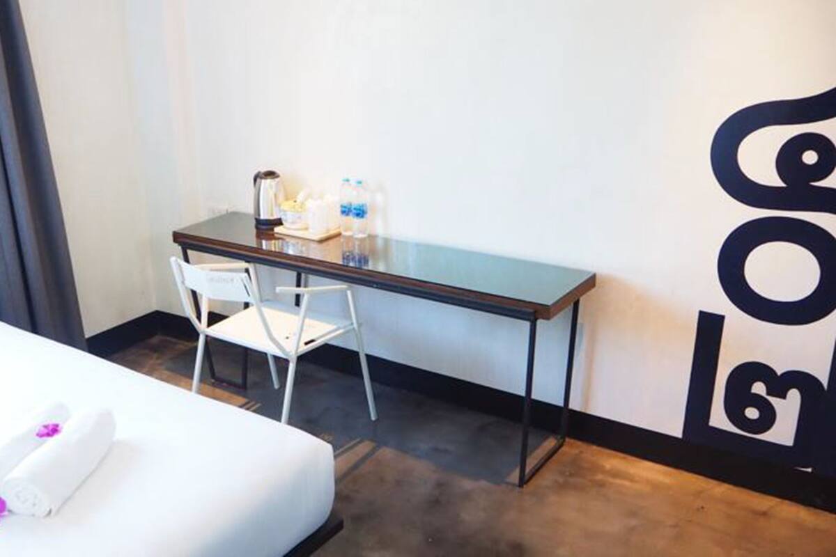 A hotel room with a bed, a desk, and a chair. There is a window with curtains, a wall with a large logo, and a small table with a lamp and a bottle of water.