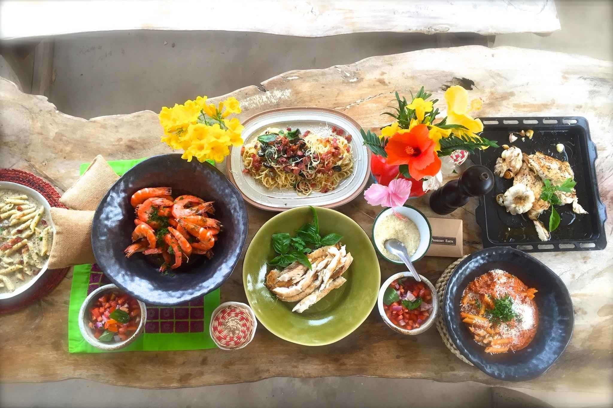 Una variedad de platos que incluyen mariscos, pasta y pollo a la parrilla, dispuestos sobre una mesa rústica con flores decorativas.