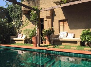 Una piscina con bordes de azulejos, un patio con mobiliario exterior y un edificio con fachada de estuco.