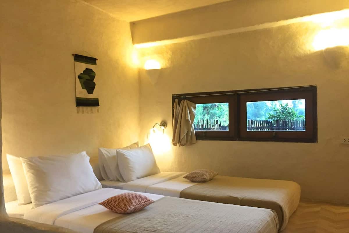 A room with two beds, a window with a view of greenery, and a wall-mounted light fixture.