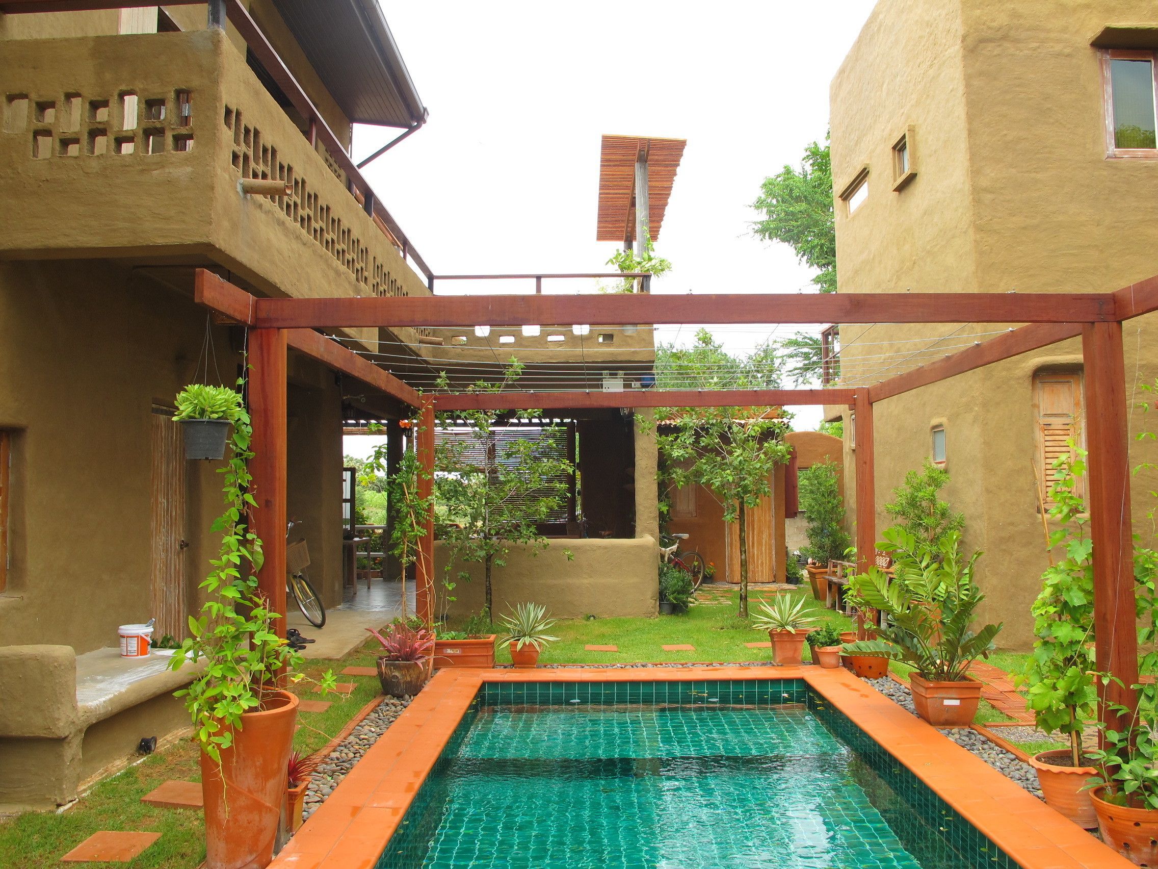 Un área de piscina con suelo de baldosas, rodeada de plantas en macetas y una pérgola.