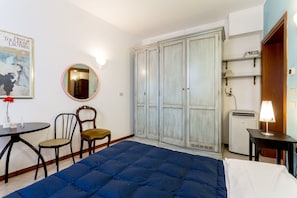 Quarto com guarda-roupa espaçoso, cama com colchão azul, espelho redondo pequeno, mesa com luminária e uma cadeira.