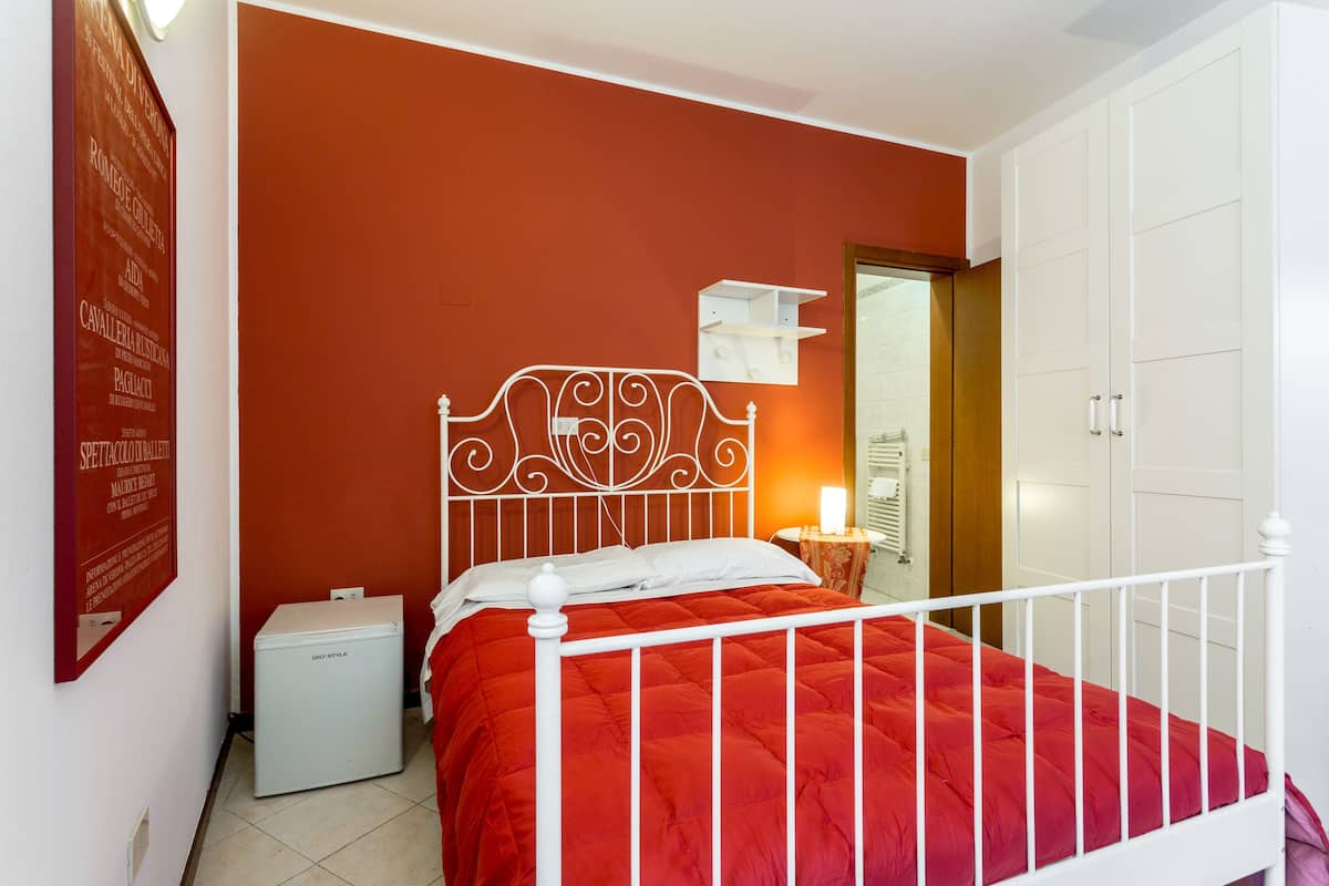 Ein Schlafzimmer mit weißem Metallbettgestell, einer roten Bettdecke, einem kleinen Nachttisch mit Lampe und einem weißen Kleiderschrank.