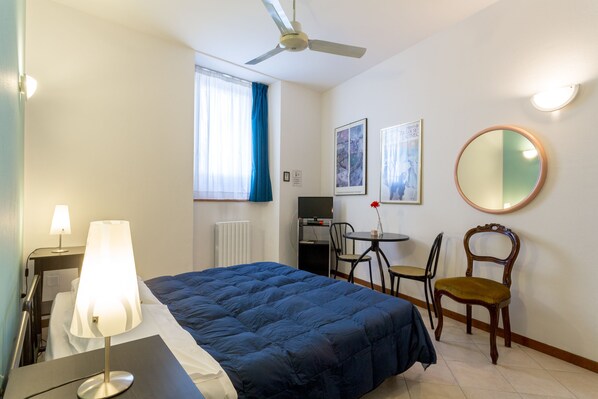 A hotel room with a bed, a desk, a chair, a round mirror, and a ceiling fan. - Alloggio Le Casette (Verona)