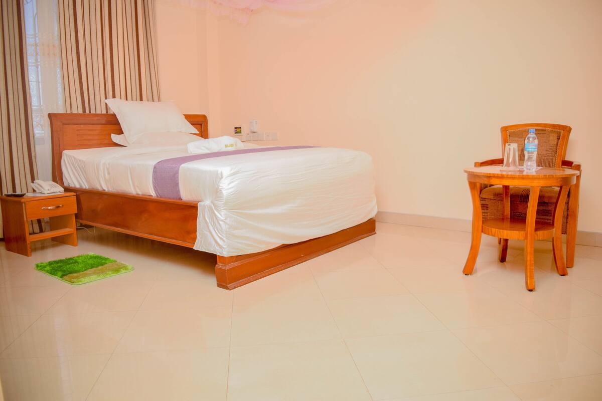 A neatly made bed with a wooden headboard, a bedside table with a phone, and a chair with a bottle on it.