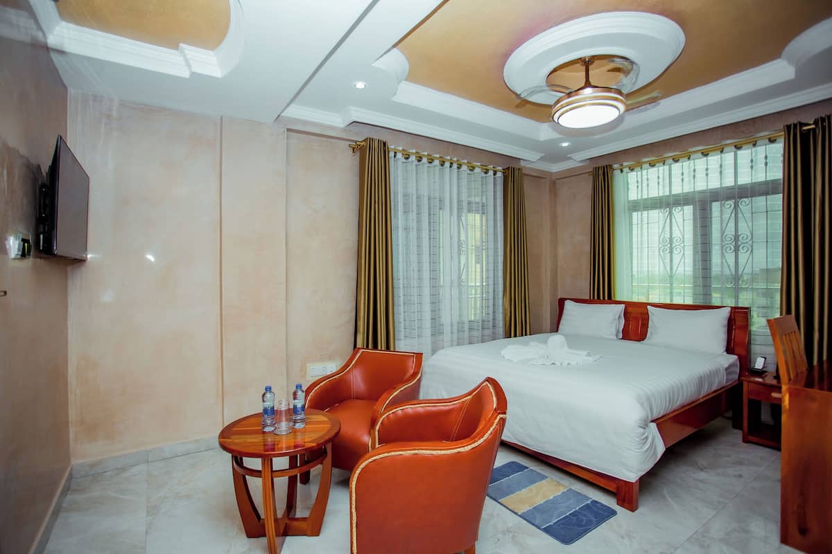 A hotel room with a large bed, two chairs, a small table with water bottles, and a TV on the wall.