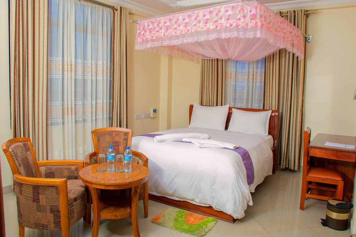 A hotel room with a bed, a desk, a chair, a small table, and a water bottle.