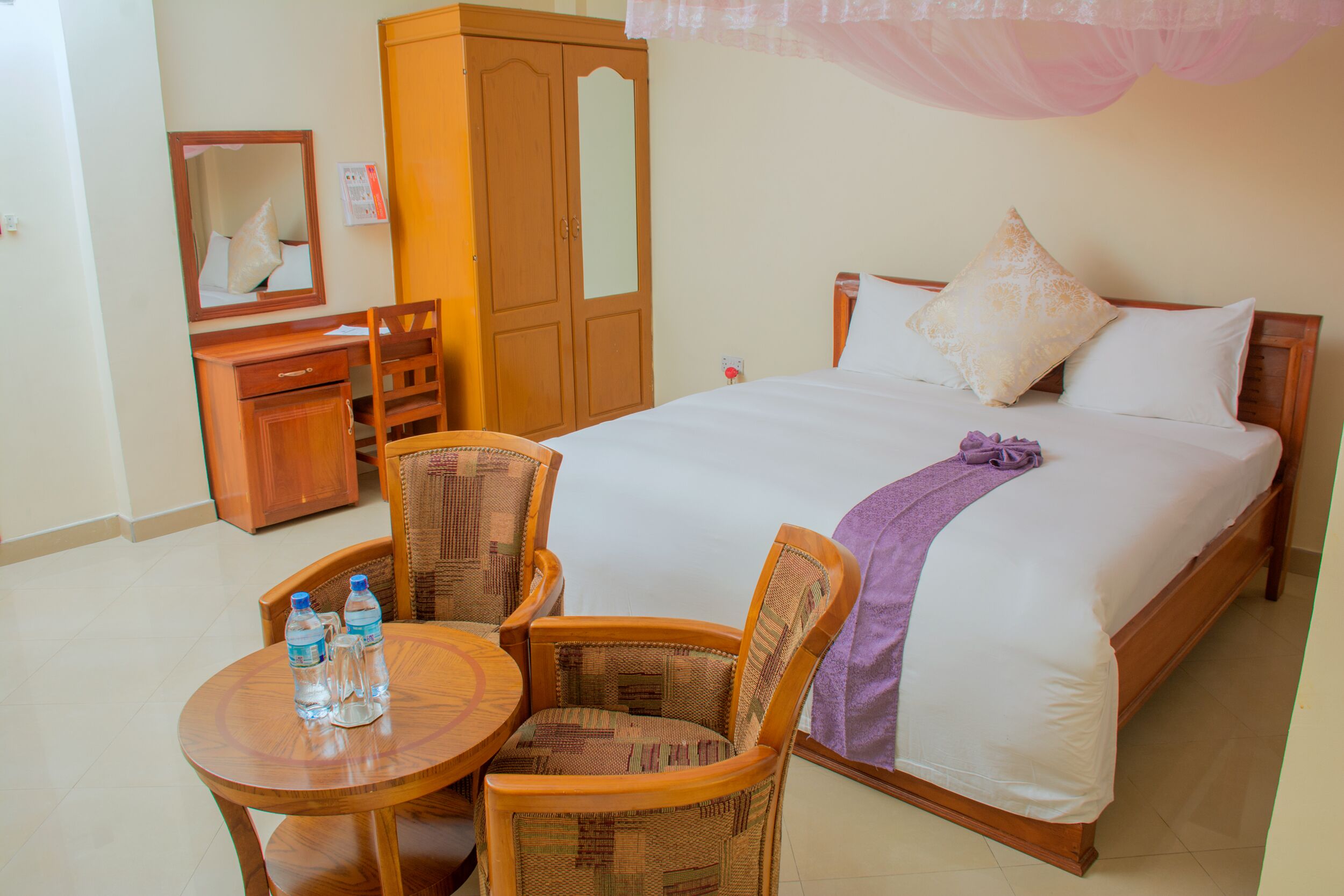 A hotel room with a bed, a wooden wardrobe, a desk with chairs, and a round table with water bottles.