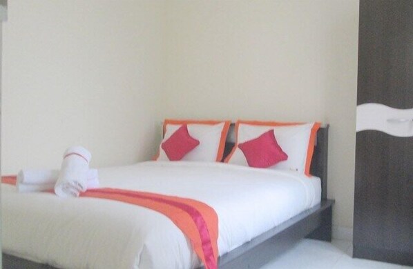 Una cama bien hecha con ropa de cama blanca y detalles en naranja, una toalla enrollada y un almohadón con estampado rojo y blanco.