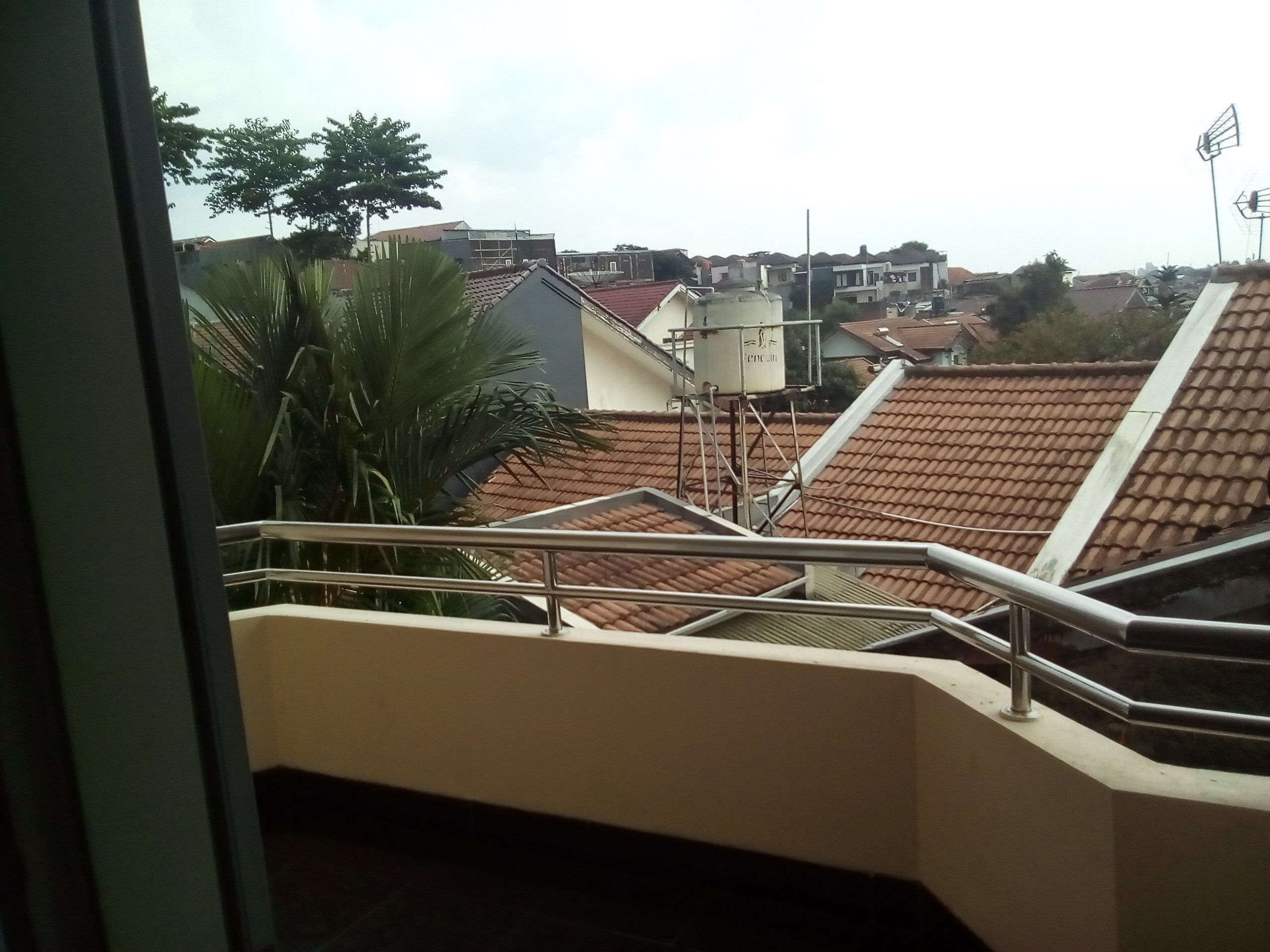 A view from a balcony with a metal railing, overlooking a residential area with tiled roofs and a water tank on a building.