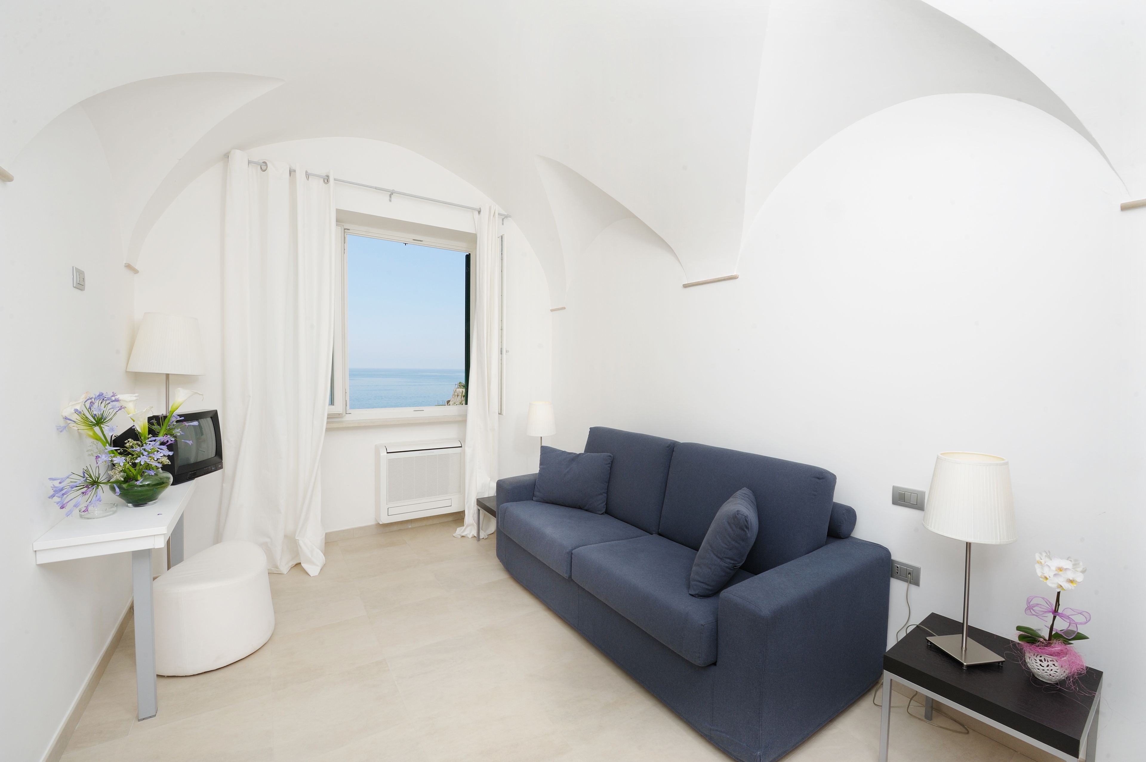 A modern living room with a blue sofa, a white desk, a TV, and a view of the sea through a window with white curtains.