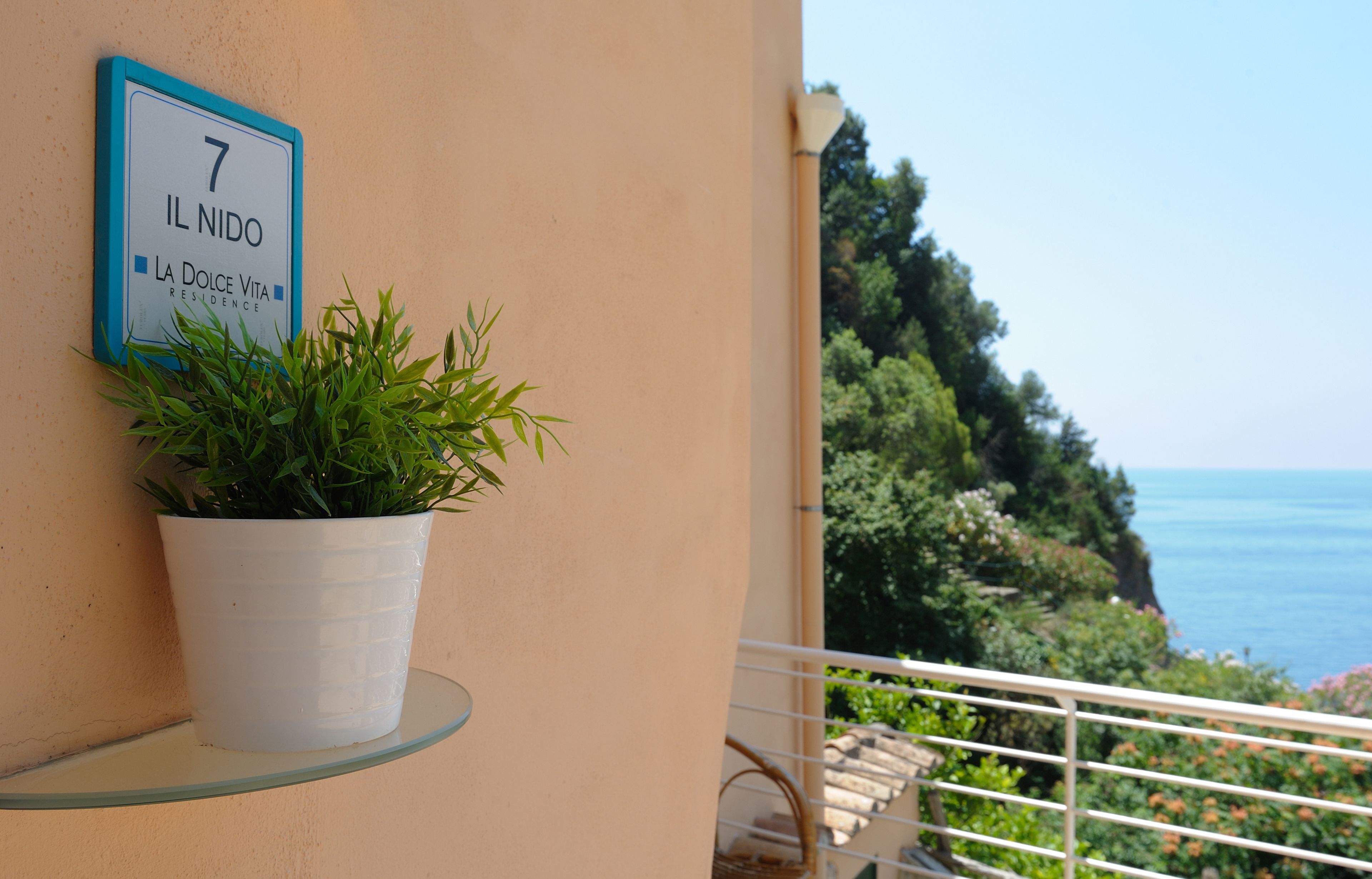 A balcony with a potted plant, a sign indicating room number 7, and a view of the sea.
