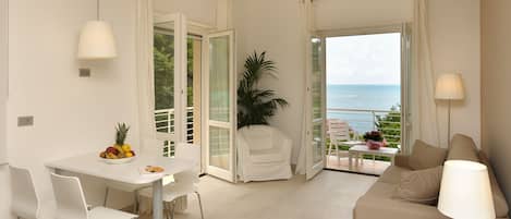 A modern living room with a dining area, a sofa, a table with fruit, and a view of the sea.