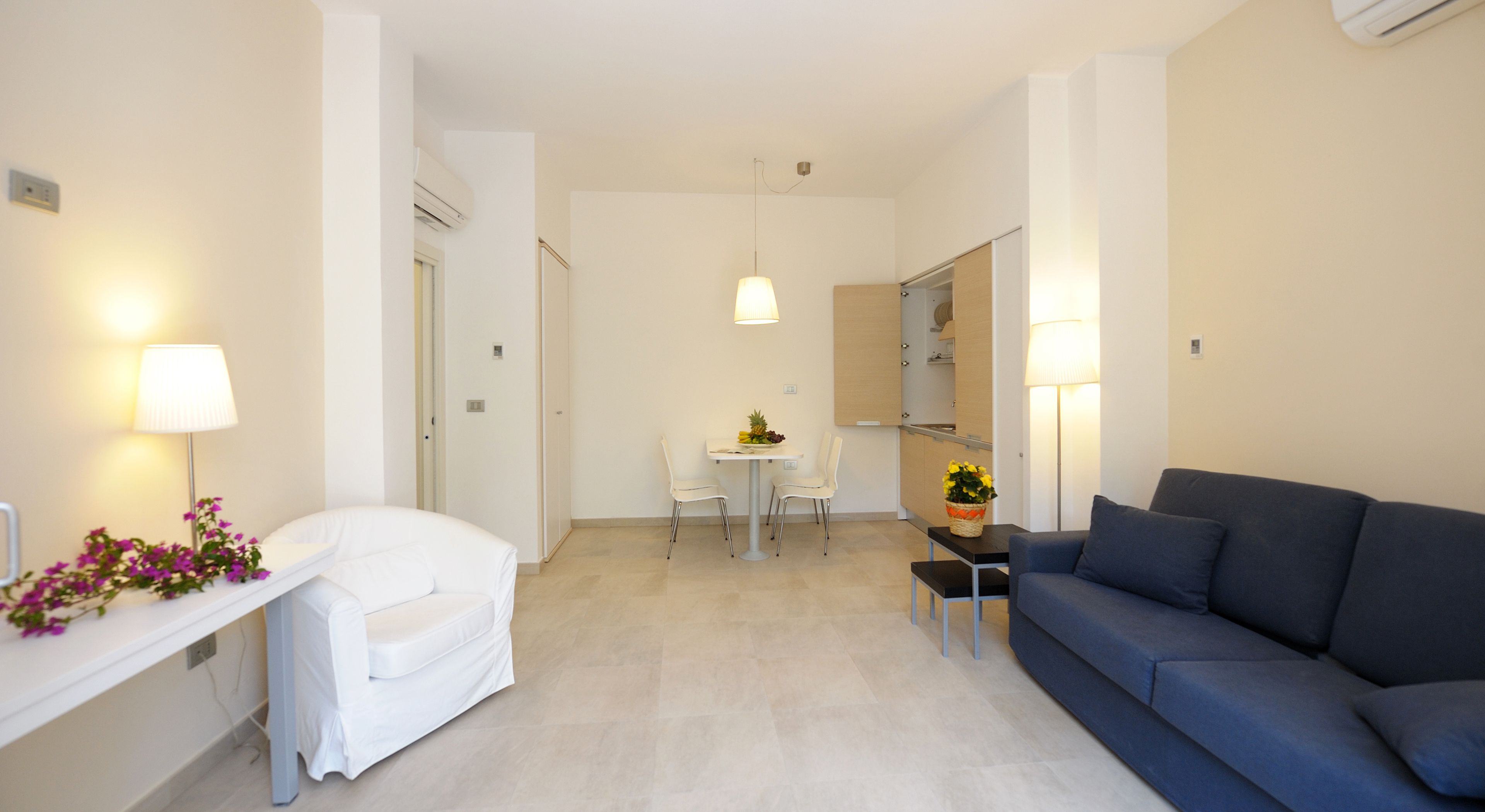 A modern living room with a sofa, a small table with a lamp, and a dining area with chairs and a table.