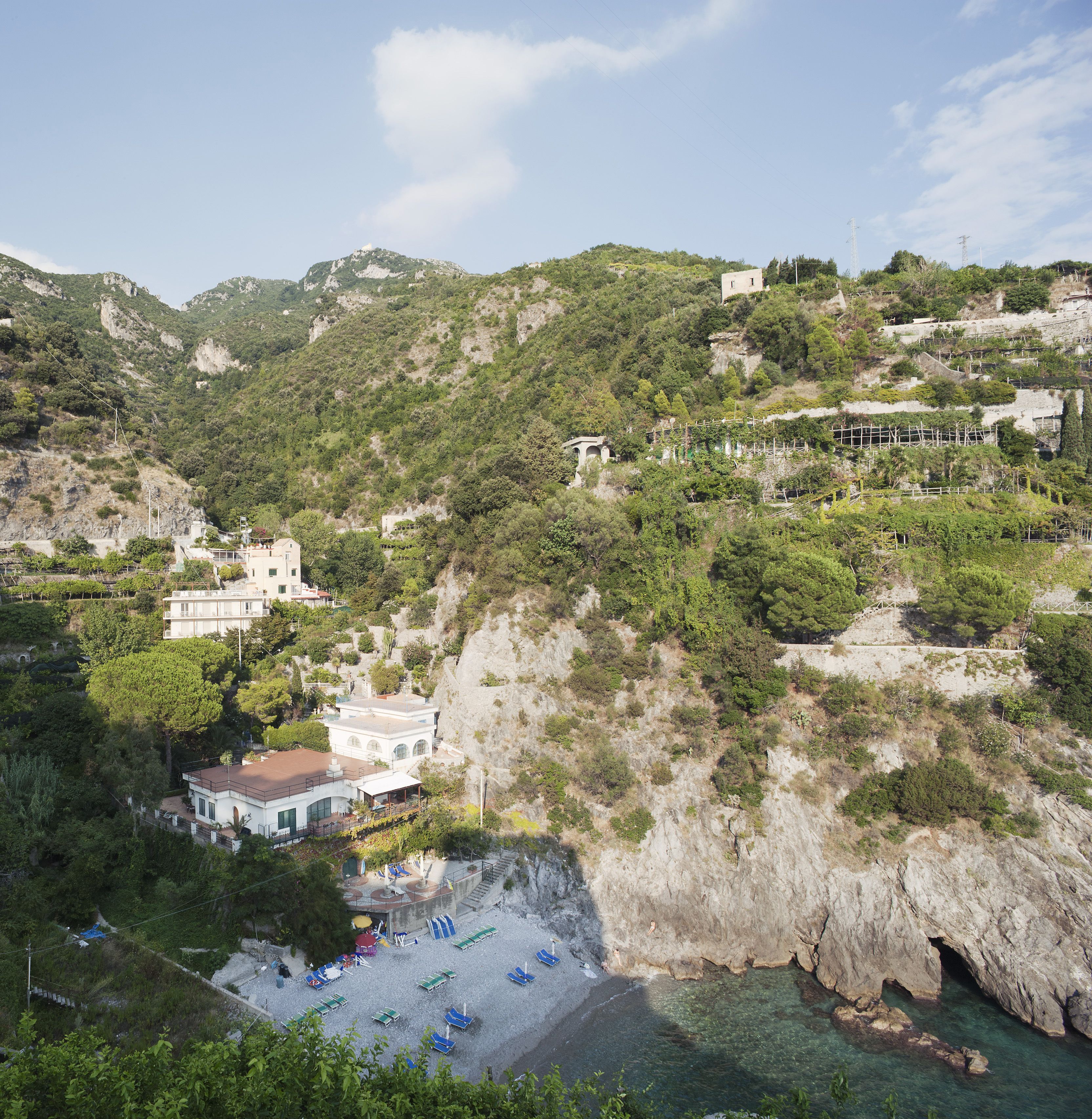 Zona costera con un promontorio rocoso, un pequeño edificio con balcón y una playa con tumbonas.