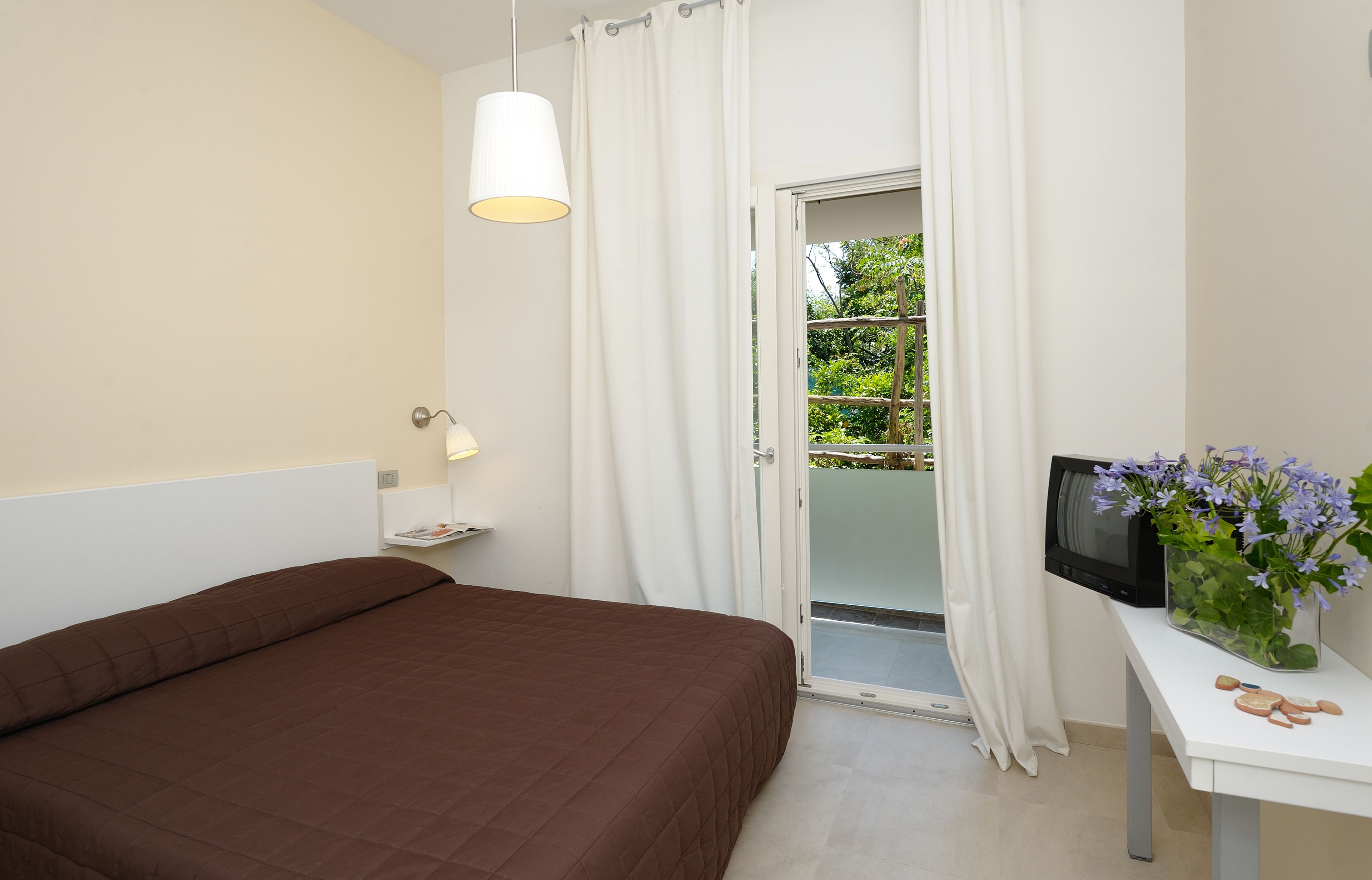 Una camera da letto con un letto, una TV, un tavolino con un vaso di fiori e una porta scorrevole in vetro che dà su un balcone.