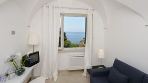 Ein Zimmer mit Fernseher, einem kleinen Tisch mit Blumenvase, einer Couch, einem Fenster mit Vorhängen und Meerblick.