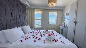 A neatly made bed with white linens and red rose petals scattered across it, a modern pendant light, and a view of the city through the window. - Buyukada Port Hotel (Adalar)